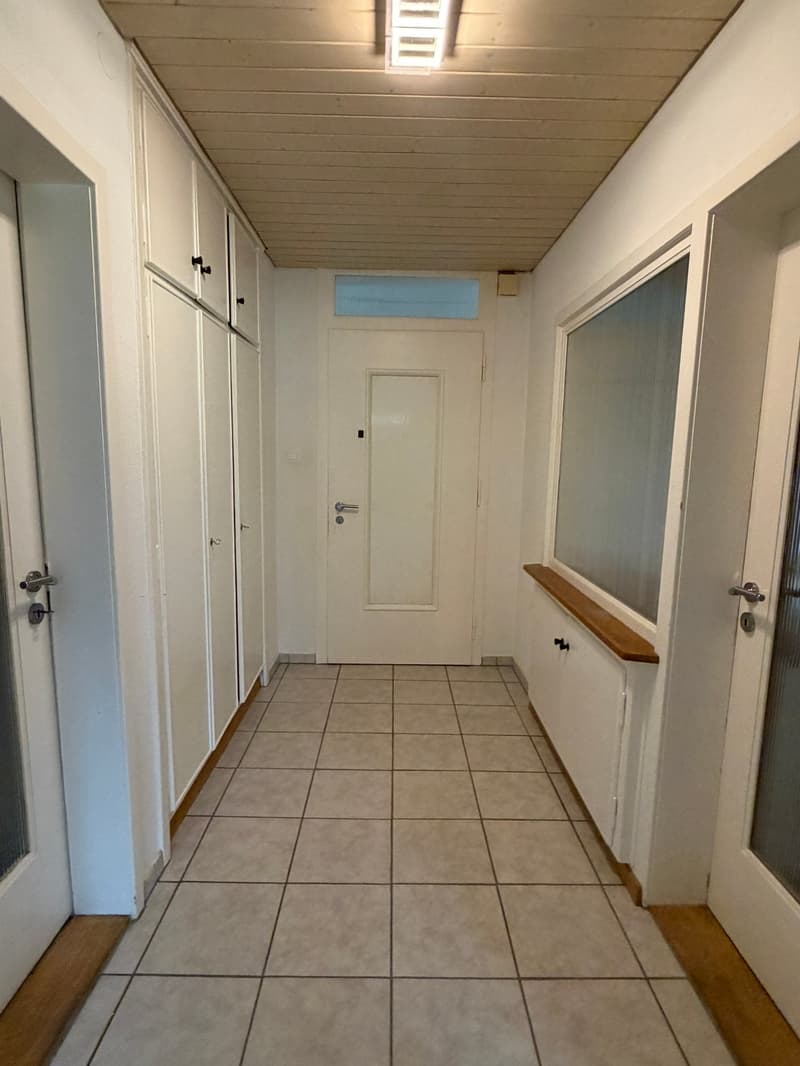 Empty hallway with tiled floor, white walls, wooden baseboards, and a white door.