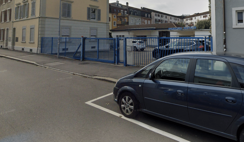 Blue parking lot with cars parked in, metal gate, buildings behind