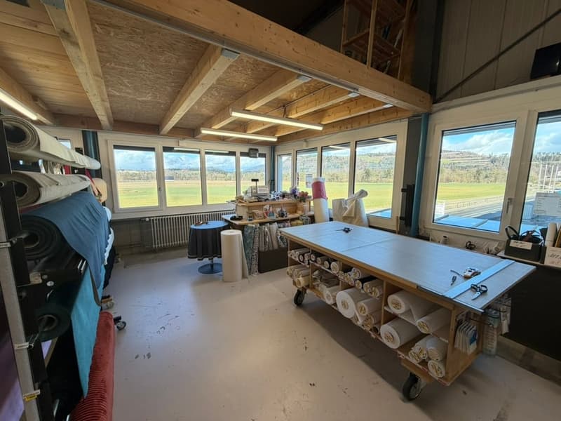 A workshop with large windows, wooden ceiling, tables, and storage racks with materials.