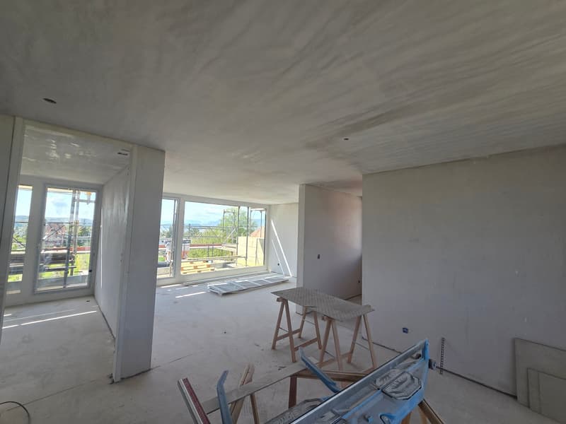 Empty room with large windows, unfinished walls, wooden workbenches, and construction materials
