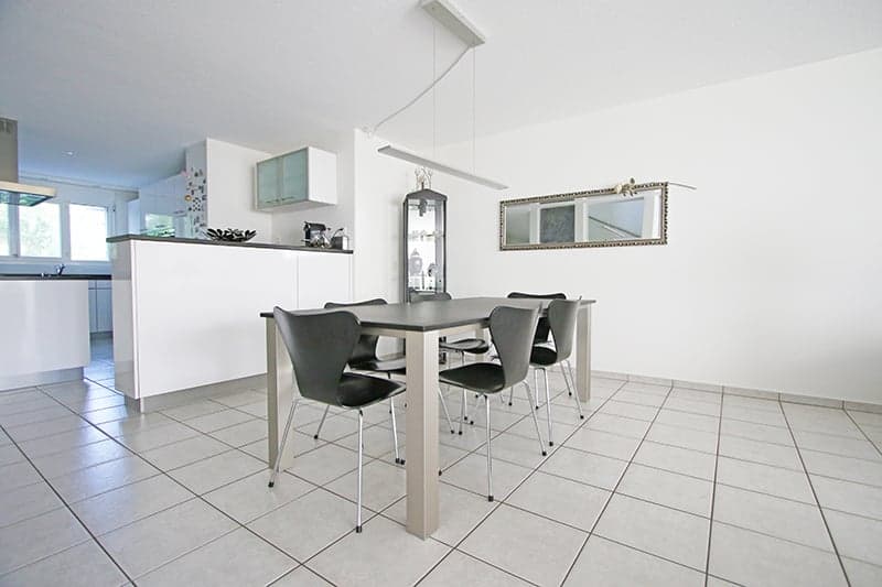 Dining area with long table, chairs, tiled flooring, modern design, counter in the background, window, kitchen island, kitchen appliances, mirror, and a lamp.