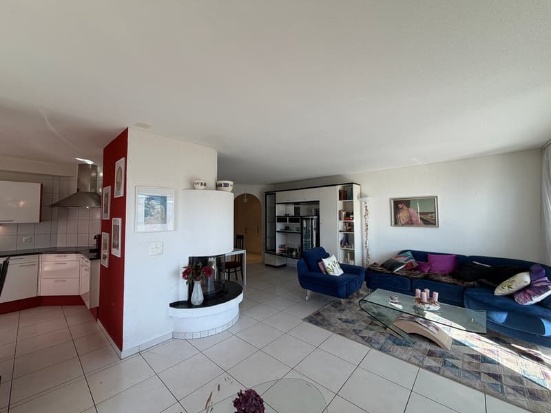 large living room with a fireplace, blue sofa, glass table, picture frames, bookshelf, red partition wall, kitchen with a countertop, cabinet, tile floor