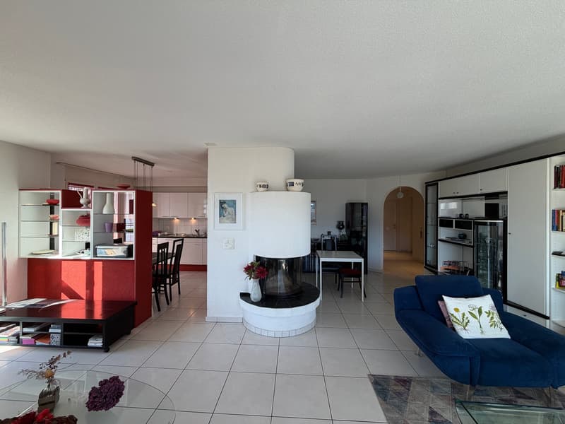 Modern living room with fireplace, blue couch, large glass table, white tiles, modern shelves, picture frame, kitchen with red wall, dining table, kitchen island, microwave, red bowl