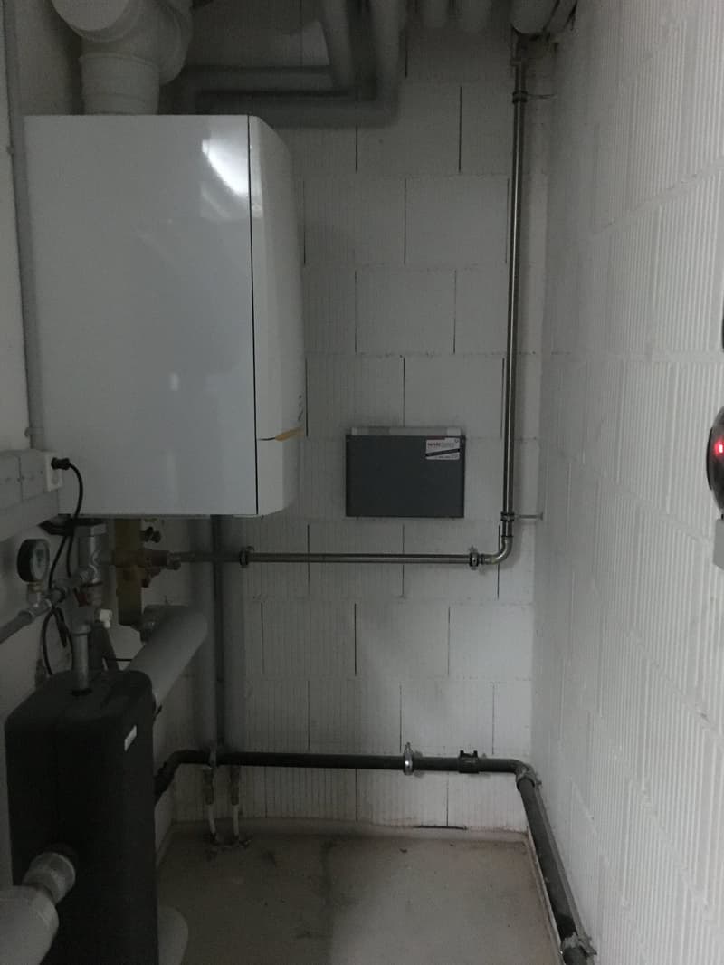 Utility room with white brick walls, white water heater, black water tank, pipes, and electrical box