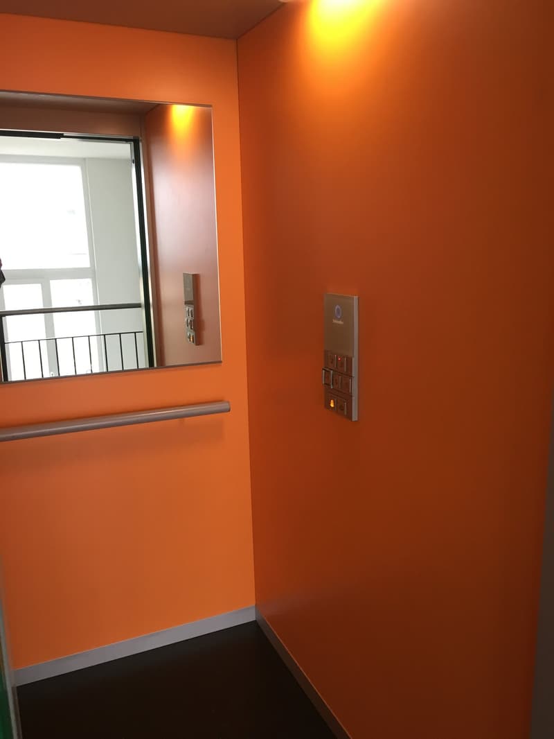 Orange wall, mirror, elevator control panel, black floor, silver handrail