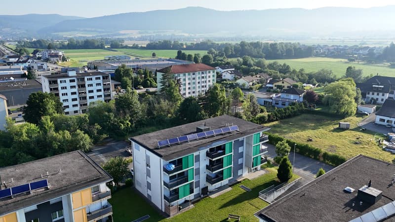 Aerial view of a residential area with multiple buildings, solar panels on the roofs, surrounded by trees and grass, with a distant view of mountains and fields.