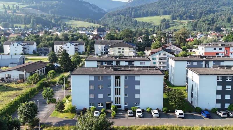 Aerial view of a residential area with multiple buildings, surrounded by greenery and mountains, with a parking lot in front of the main building.