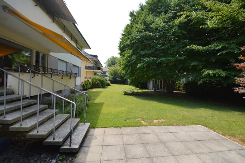 building with stairs leading to a well-maintained lawn, yellow awning, trees, a small shed, and another building