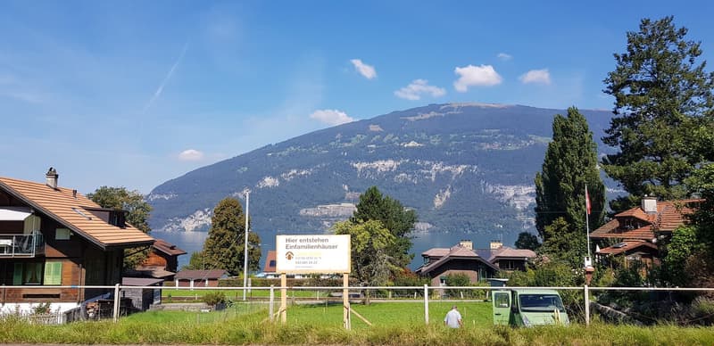 The image shows a scenic landscape with a lake, mountains, and traditional Swiss-style houses. There is a sign advertising new family homes. The scene appears to be in a rural or small town setting with lush greenery and a peaceful atmosphere.