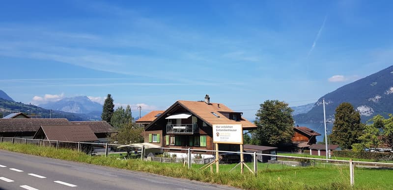 Two-story wooden chalet-style house with a red tile roof, surrounded by mountains and greenery. The house has balconies on the upper floor and is located next to a road with a sign indicating the construction of new residential buildings.