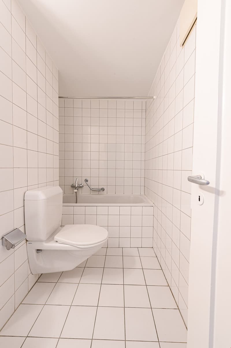All white tiles, corner toilet, bathtub with grab bar, closed door