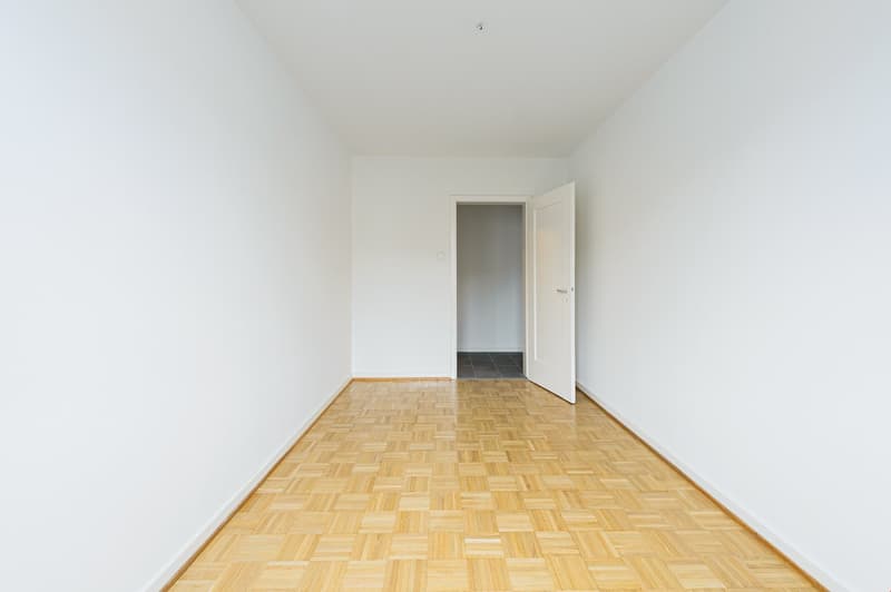 Empty room with wooden floor, white walls, open door leading to another room.