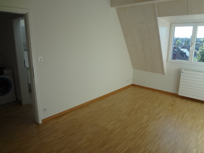 Empty room with wooden floors, white walls, a window on the right, and an open door leading to a corridor with a washing machine.