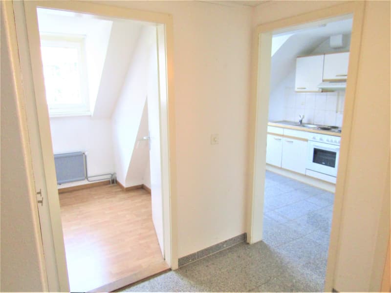 Empty room with wooden floors and slanted ceiling, leading to a kitchen with a stove and sink.