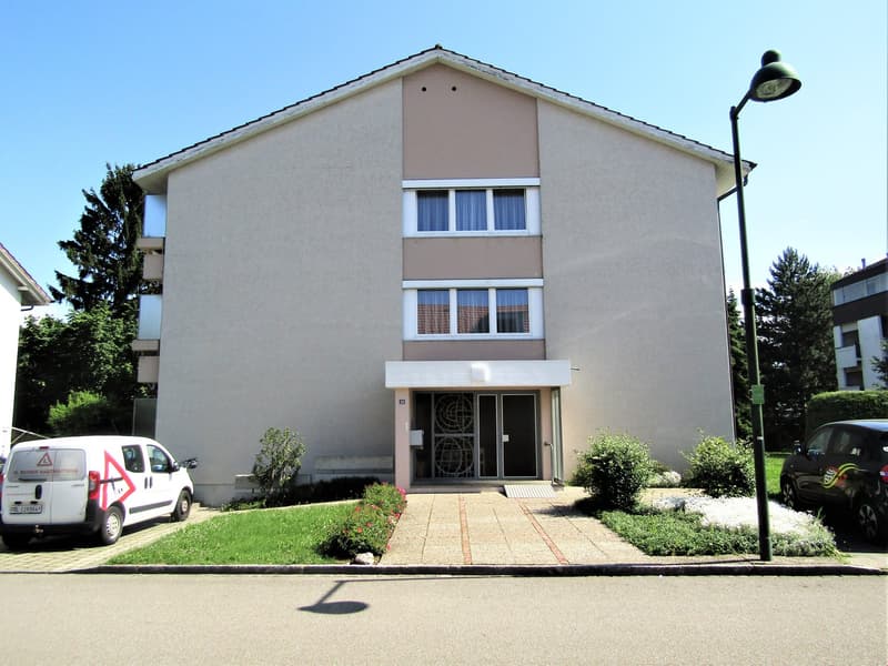 2 story apartment building, beige exterior, windows with white frames, entrance with double doors, paved walkway, two cars parked in front