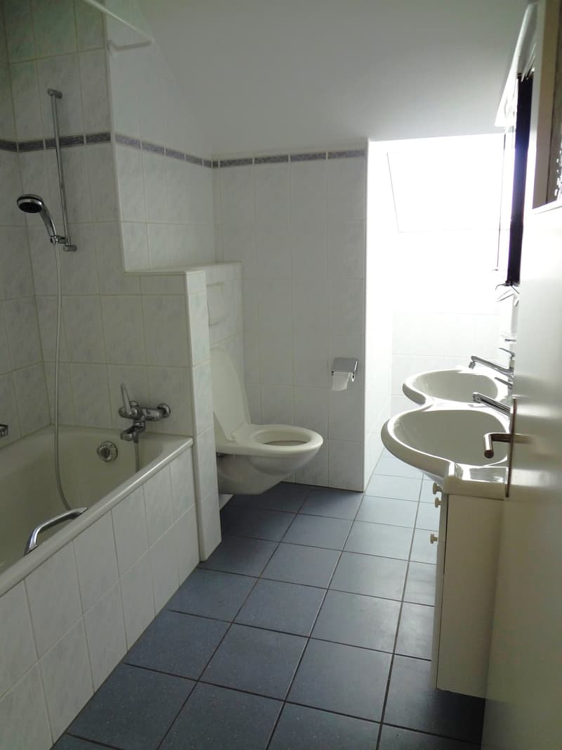 The image shows a bathroom with white tiled walls, a bathtub, a toilet, and a double sink vanity. The floor is covered with dark gray tiles. The bathroom appears to be clean and well-maintained.