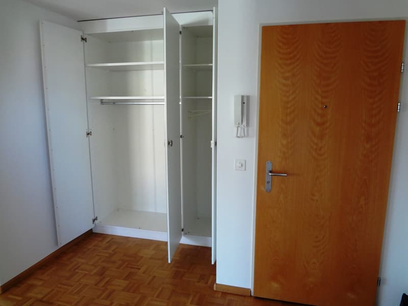 The image shows a bedroom with a built-in wardrobe, hardwood flooring, and a wooden door. The wardrobe has multiple shelves and hanging space. The room appears to be simple and functional.