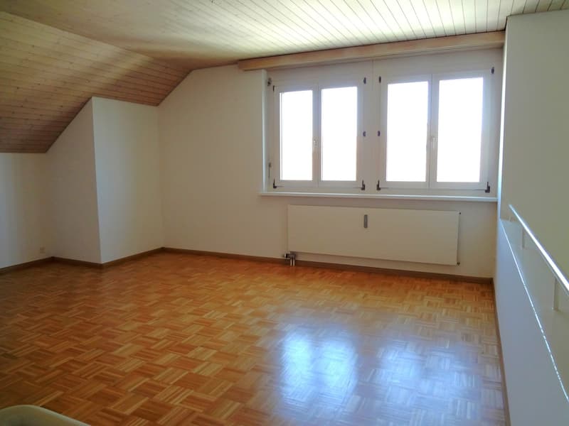 Spacious bedroom with wooden floor, white walls, and large windows providing ample natural light. The room has a slanted ceiling with wooden paneling.