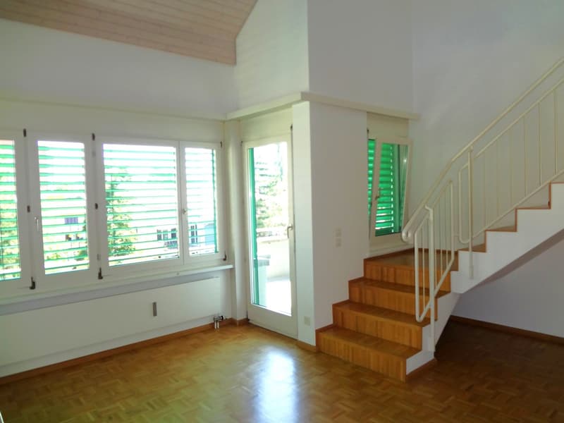 Spacious room with large windows, wooden floors, and a staircase leading to an upper level. The room has a bright and airy feel with natural light coming in through the windows.
