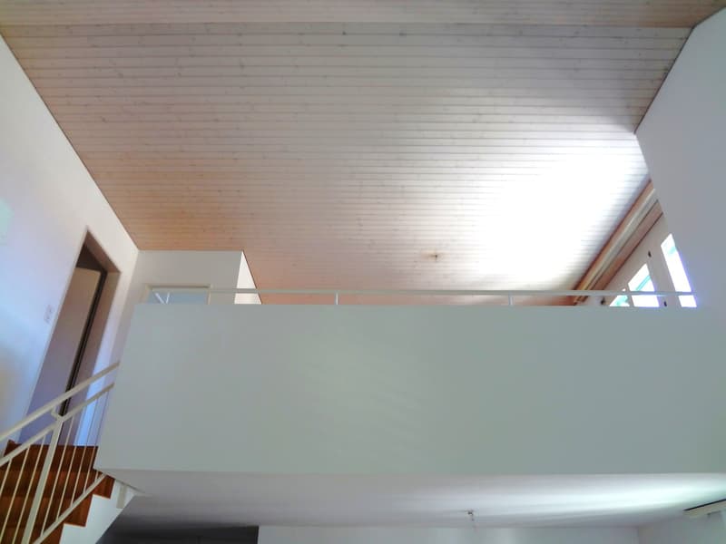 The image shows a room with a high ceiling and wooden paneled walls. There is a balcony or terrace visible through the window, suggesting this is a living or dining area with access to an outdoor space.