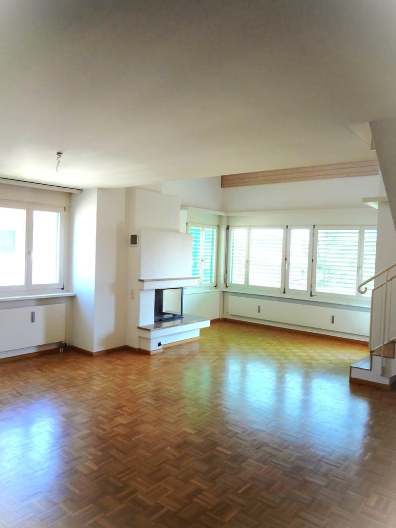 Spacious living room with hardwood flooring, large windows, and a fireplace. The room has a clean, minimalist design with white walls and wooden accents.