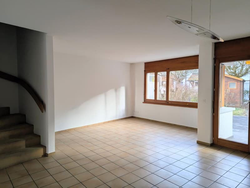 Empty room with white walls, tiled floor, wooden window frames, and a staircase on the left.