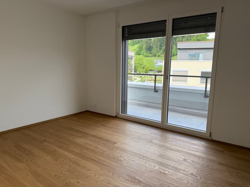 Spacious room with hardwood flooring, large windows providing a view of the outdoor greenery, and a sliding glass door leading to a balcony or terrace.