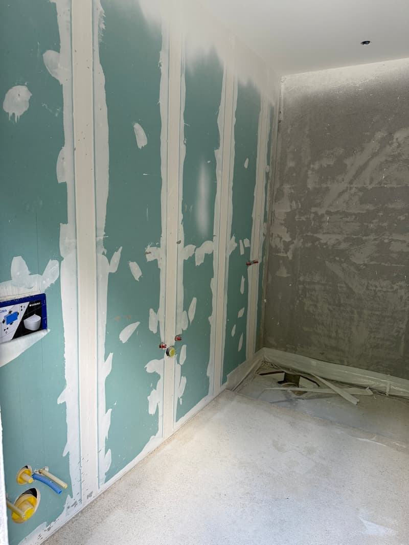 The image shows a room undergoing renovation. The walls are partially covered with drywall, and the floor is covered in construction materials. There are tools and supplies visible, indicating that the room is in the process of being remodeled or repaired
