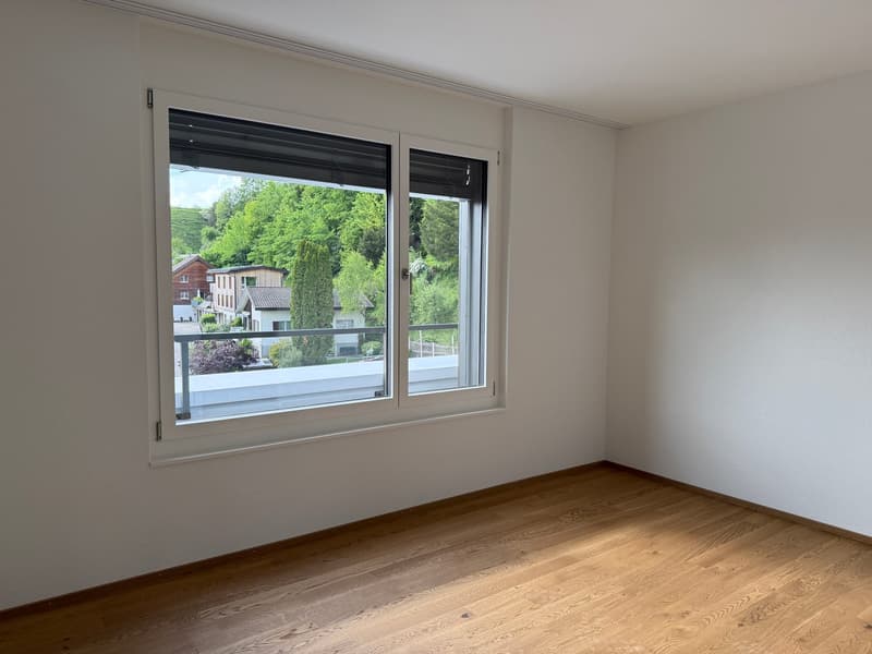 Spacious bedroom with large windows overlooking a scenic outdoor view of trees and buildings. The room has hardwood flooring and white walls.