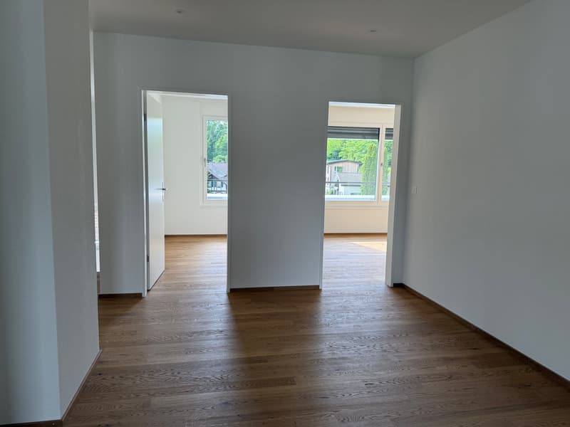 This image shows an empty room with white walls and hardwood flooring. The room has two large windows that provide natural light and views of the surrounding area. The room appears to be a living room or bedroom, with an open layout and minimal furnishing