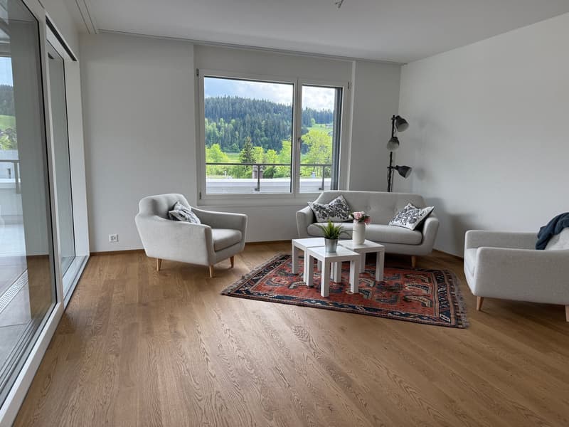 Spacious living room with large windows offering a scenic view of the surrounding forest and landscape. The room features a gray sofa, armchair, and coffee table on a patterned rug. The hardwood flooring and neutral wall colors create a modern and minimal