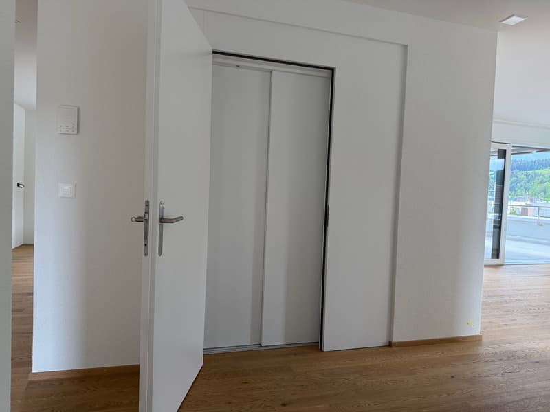 The image shows an empty room with white walls, a wooden floor, and a sliding door closet. The room appears to be a bedroom or living space with a simple, minimalist design.