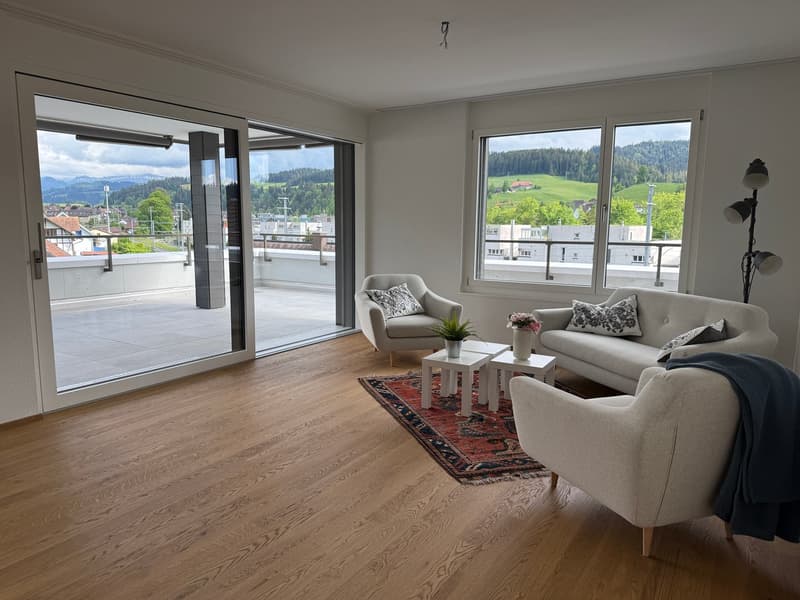 Spacious living room with large windows offering a scenic view of the surrounding countryside. The room features hardwood flooring, a comfortable sofa and armchair, and a decorative rug. The room has access to a balcony or terrace through sliding glass do