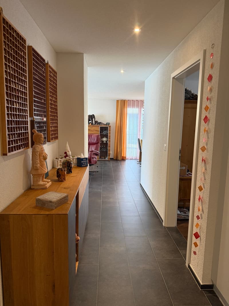 gray tiled floor, long hallway, wooden cabinet with items on top, wooden display shelves on the wall, bright lighting, hallway leads to a room with an open door