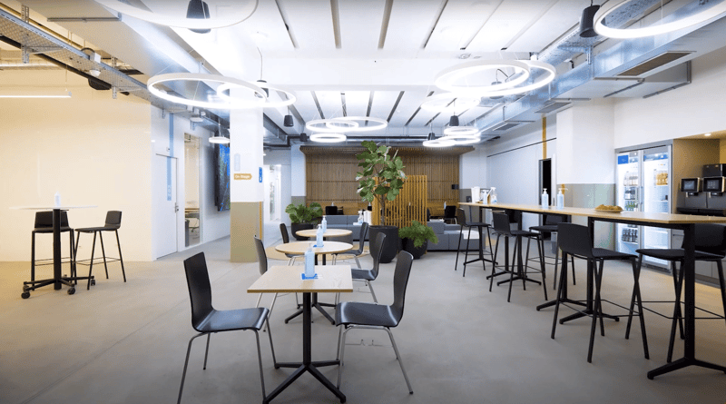 open plan office with tables, chairs, potted plants, dispenser, refrigerator, ceiling lights
