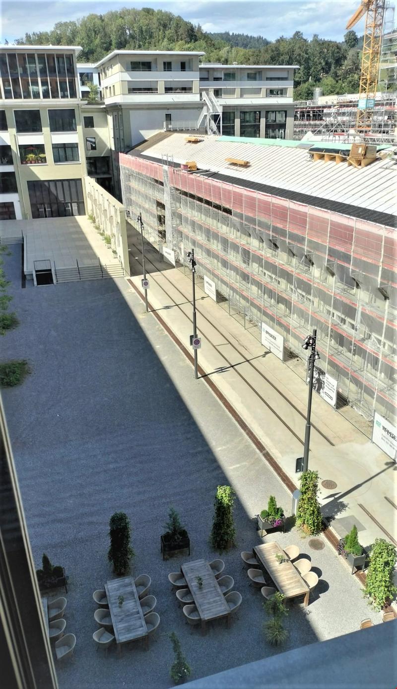 an outdoor area with several buildings, under construction. There are street lights, a building covered in scaffolding, and a parking lot with a row of tables and chairs.