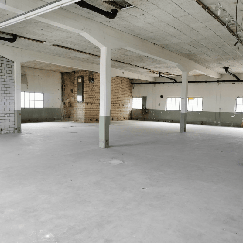 Large open space with concrete floors, exposed brick walls, and a high ceiling with visible ductwork and pipes. The space appears to be unfinished and in need of renovation.