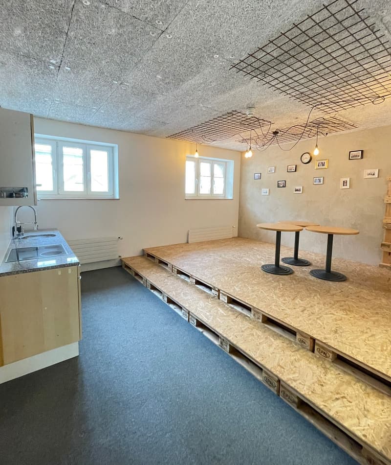 empty room, elevated platform made of wooden pallets, kitchen area with sink and faucet, white walls, wooden ceiling, multiple windows, square frames hanging on the wall