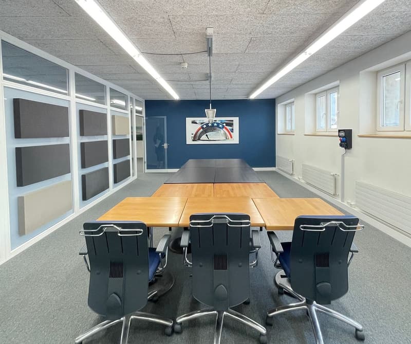 Empty room with long tables and black swivel chairs, gray carpet, white and blue walls, lamps on ceiling, large window, picture on wall, heating elements