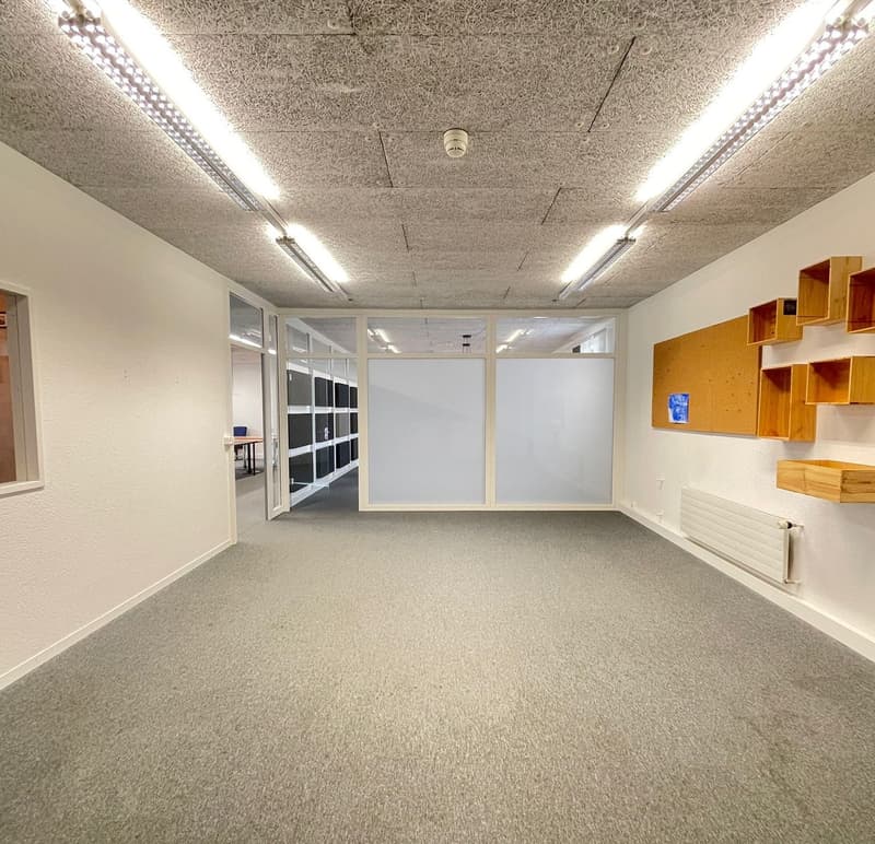 This image shows a large open office space with a carpeted floor, white walls, and fluorescent lighting fixtures on the ceiling. The room has a glass partition wall that separates it from another area. There are some built-in shelves or storage units alon
