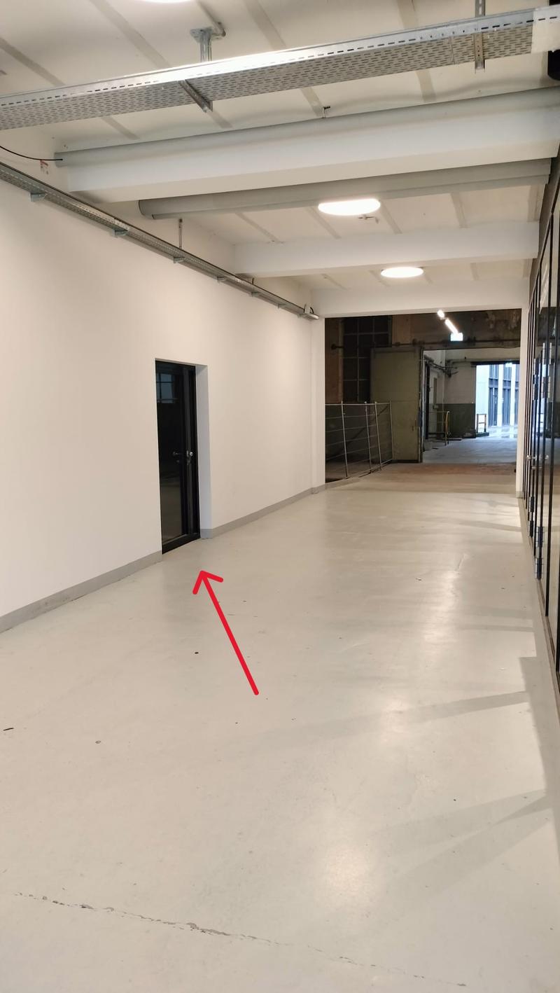The image showcases a large, vacant room with industrial design elements. It has exposed ceiling beams, metal shelving, and bright fluorescent ceiling lights. The floor is polished concrete. The room is divided into sections with metal partitions. One sec