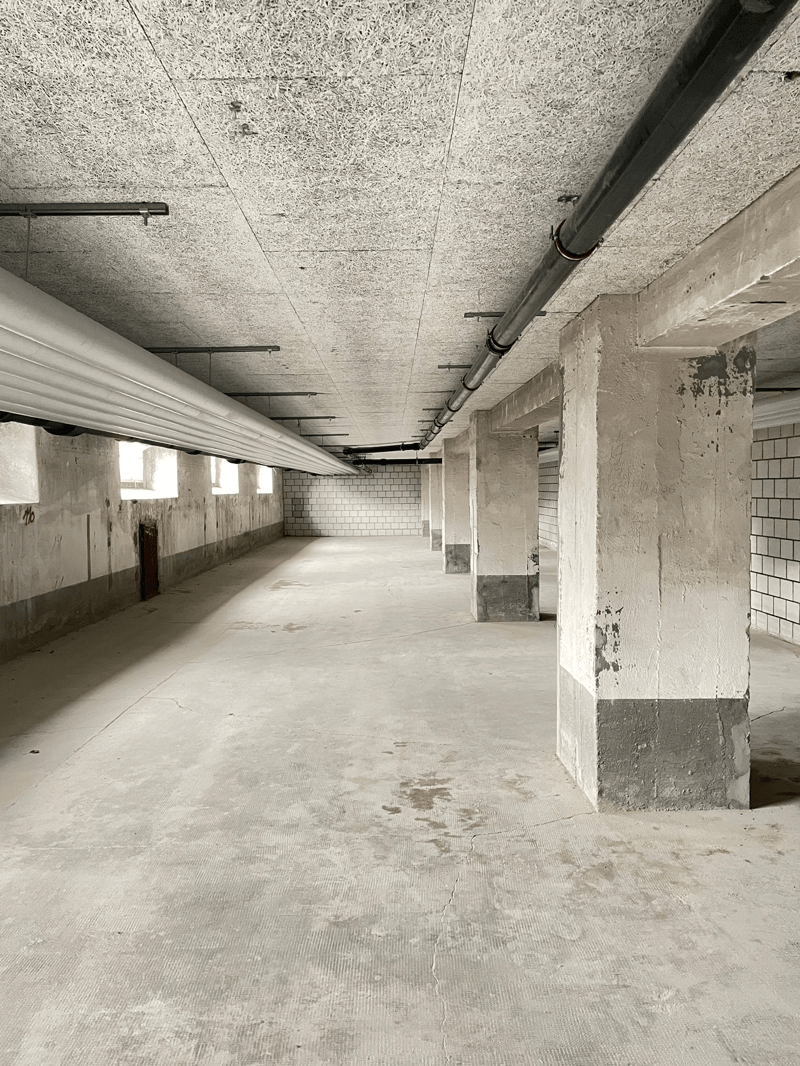 Empty room, white brick walls, white ceiling, gray floor, several columns, pipes along the ceiling and floor, two open windows on one wall, dark mark on the wall