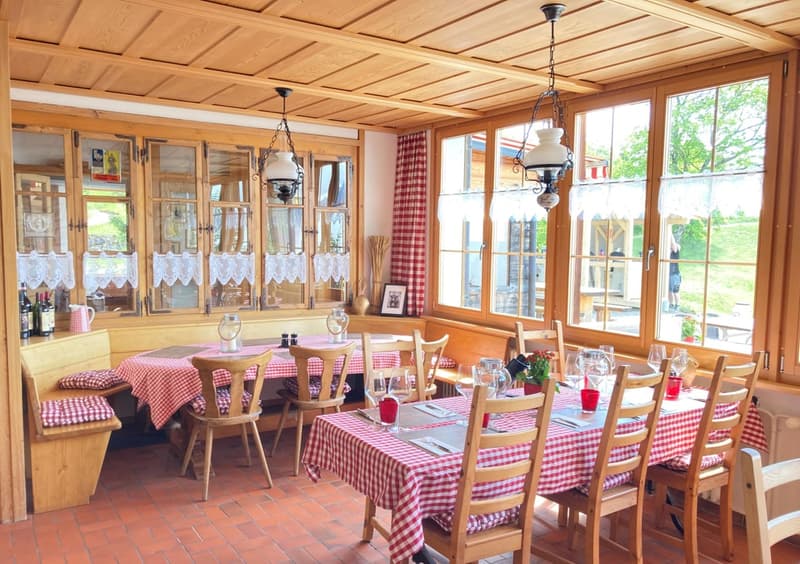 wooden paneling, wooden floor, dining table with chairs, checkered tablecloth, wine glasses, forks, spoons, knives, plates, two lamps, windows, view to the outside