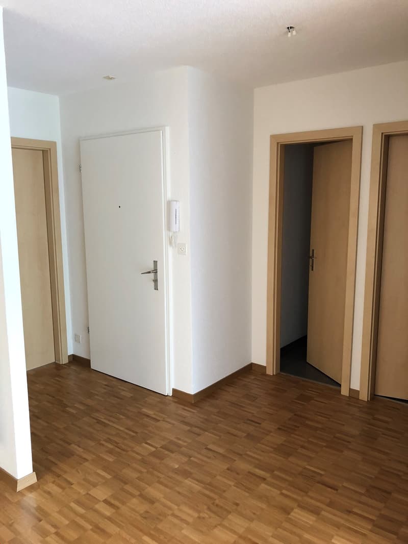 Empty room with wooden floors, white walls, three doors, one with a doorbell, and a power outlet.