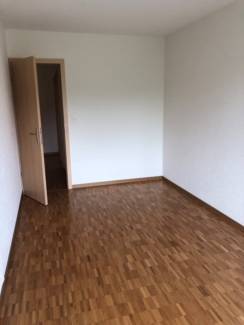 Empty room with wooden floor, white walls, open door leading to another room, power outlet on the wall