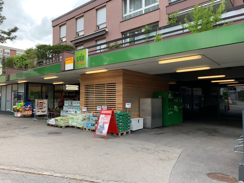 The image shows the exterior of a retail store called 'Uhi Anchors'. The store has a green and orange color scheme, with a covered entrance area and parking spaces in front. There are various products and displays visible outside the store, including bags
