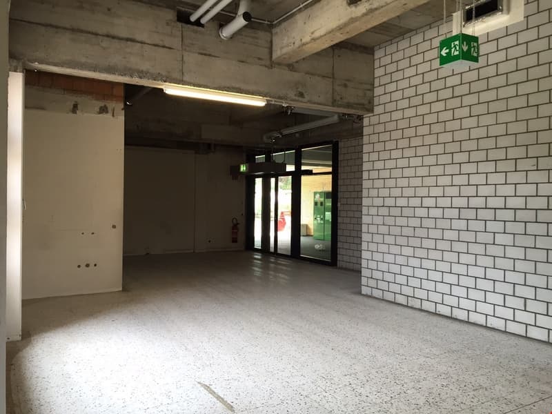 The image shows a large, open commercial space with concrete walls, a tiled floor, and exposed ceiling pipes. The space has a green exit sign and appears to be an unfinished or partially renovated area, possibly a retail or office space.