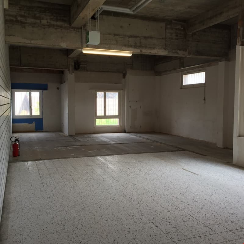 This image shows a large, open commercial space with concrete floors, exposed ceiling beams, and several windows. The space appears to be unfinished or under renovation, with a bare, industrial feel. There is a fire extinguisher visible, indicating this c