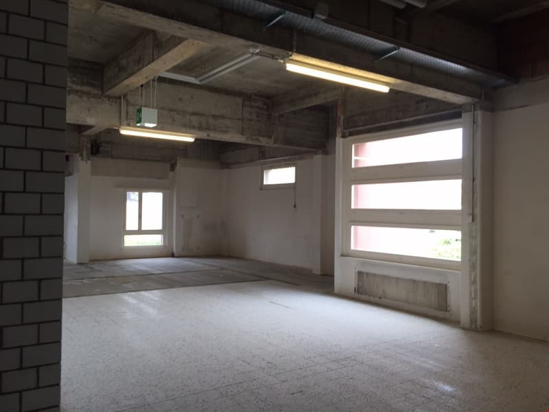 The image shows a large, open commercial space with concrete floors, exposed brick walls, and overhead lighting. The space appears to be unfinished or under renovation, with a few windows providing natural light. The overall impression is of a raw, indust
