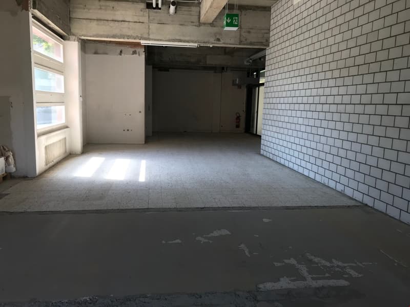 This image shows a large, empty room with concrete floors and white tiled walls. The room appears to be in a state of renovation or construction, with exposed pipes and wiring visible on the ceiling. There is a window on one side of the room, allowing nat